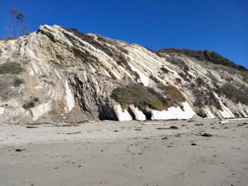 A sandy beach with a steep, rocky cliff and clear blue sky in the background.