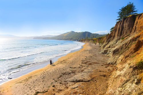 A scenic beach with cliffs, gentle waves, and people walking along the shore under a clear blue sky.