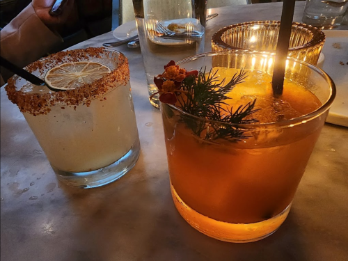 Two cocktails on a table: one with a salted rim and lemon slice, the other garnished with herbs and flowers.