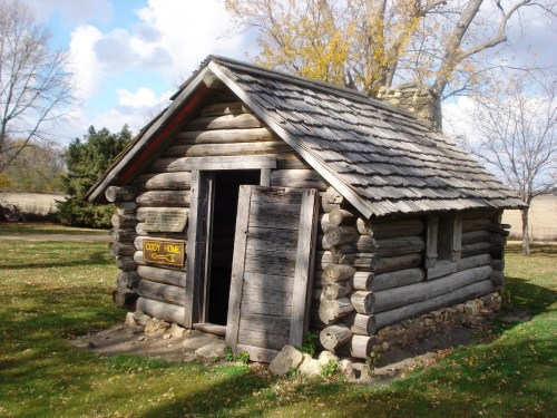 A rustic log cabin with a shingled roof, set in a grassy area with trees in the background.