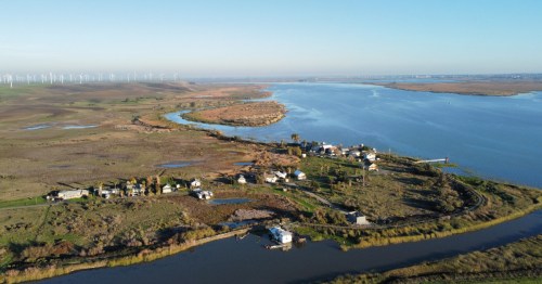 Aerial view of a river winding through a landscape with houses, fields, and wind turbines in the distance.