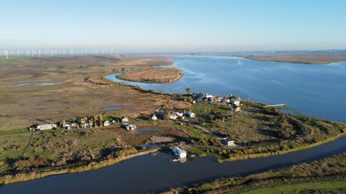 Aerial view of a serene landscape with a river, winding paths, and small houses surrounded by greenery and wind turbines.