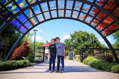 Two children stand under a colorful archway in a park, surrounded by greenery and a bright blue sky.