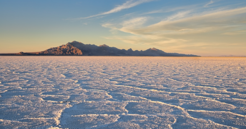 A vast salt flat stretches towards distant mountains under a colorful sky at sunset.