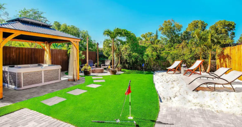 A sunny backyard featuring a hot tub, lounge chairs, putting green, and sandy area surrounded by lush greenery.