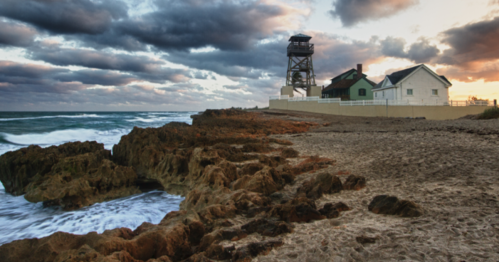 A rocky beach at sunset with a lighthouse and a house in the background, under a cloudy sky.