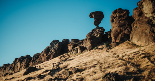 A person walks along a rocky hillside with a unique rock formation against a clear blue sky.