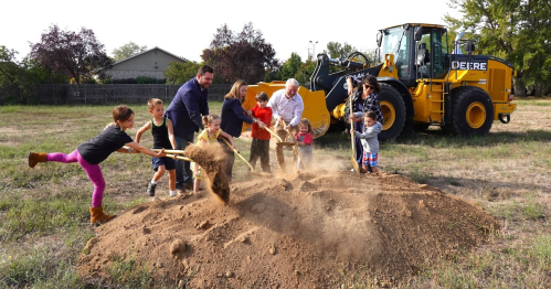 A group of adults and children joyfully shovel dirt at a construction site, with a large yellow bulldozer in the background.