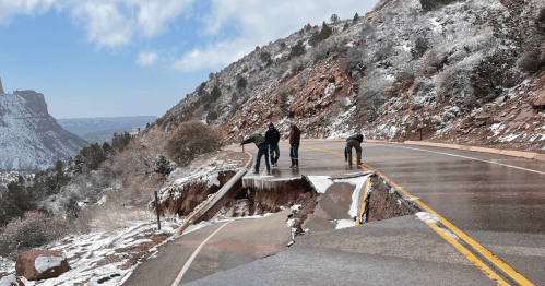 Four people inspect a damaged road on a snowy mountain, with a significant section eroded away.