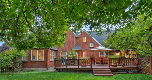 A cozy backyard with a wooden deck, patio furniture, and lush greenery surrounding a charming brick house.