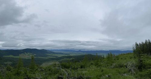 A panoramic view of a lush green landscape under a cloudy sky, with rolling hills and distant mountains.