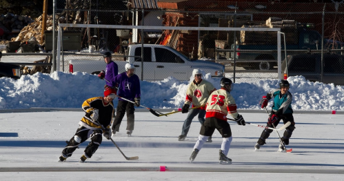 A group of six people playing ice hockey on a snowy outdoor rink, with a truck and logs in the background.
