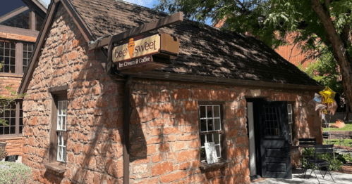 A quaint stone building with a sign reading "Beesweet by Cream & Coffee," surrounded by trees and outdoor seating.