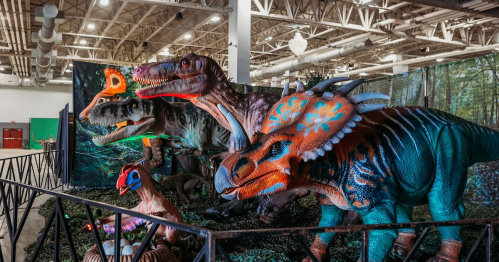 A display of colorful dinosaur models in a museum-like setting, surrounded by greenery and artificial scenery.