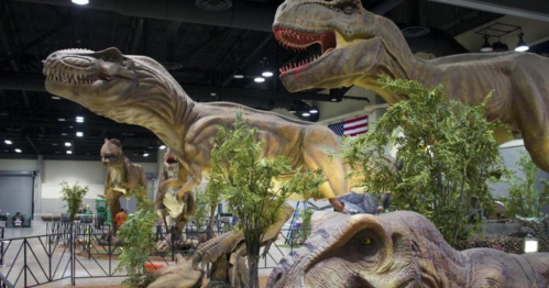 A display of life-sized dinosaur models in a convention center, surrounded by greenery and exhibit structures.