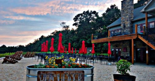 Outdoor seating area with red umbrellas, flowers, and a stone building under a colorful sky at sunset.