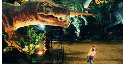 A child holds a baby while standing near a large dinosaur exhibit in a dimly lit environment.