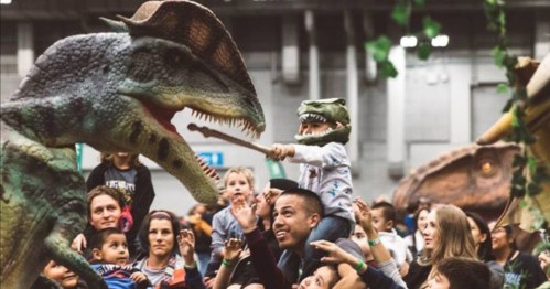 A crowd enjoys a dinosaur exhibit, with a child playfully interacting with a large dinosaur puppet.