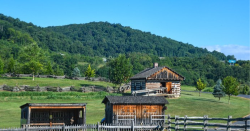 A rustic log cabin surrounded by green hills and trees, with a wooden fence in the foreground. Clear blue sky above.