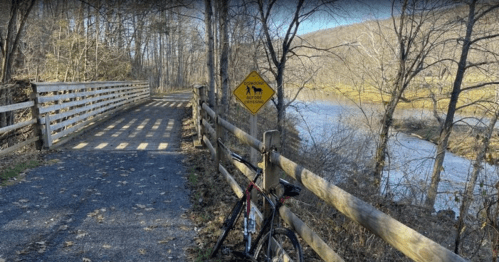 A bike rests against a wooden fence near a river, with a warning sign for pedestrians and horses on a scenic trail.