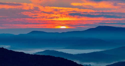 A vibrant sunset casts orange and pink hues over misty blue mountains and valleys.