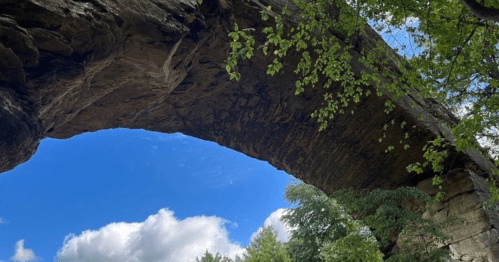 A natural stone arch surrounded by greenery under a blue sky with fluffy clouds.