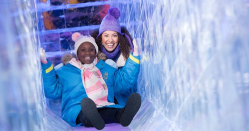 Two smiling women slide down an icy tunnel, wearing winter coats and hats, surrounded by colorful ice sculptures.