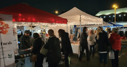A bustling night market scene with vendors under tents, string lights, and shoppers exploring various stalls.