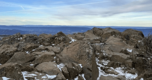 Rocky mountain summit with snow patches, overlooking a vast valley and distant mountains under a cloudy sky.