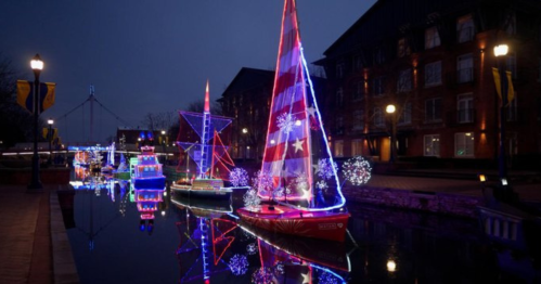 Colorful illuminated boats with festive lights reflect in a calm waterway at night, surrounded by buildings.