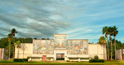 A large mural depicting various scenes, set on a building surrounded by palm trees under a cloudy sky.
