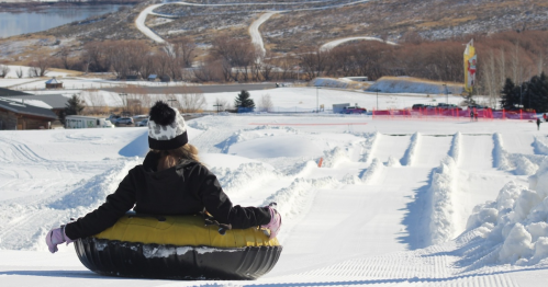 A person in a black jacket and hat sits on a yellow snow tube, sliding down a snowy hill with a scenic background.