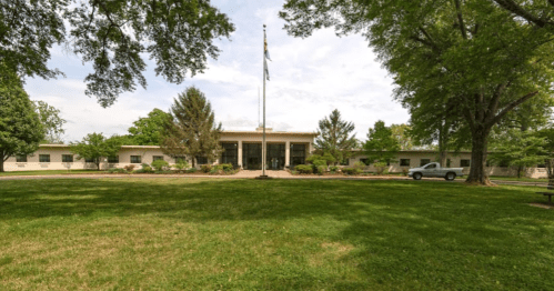 A modern building with a flagpole, surrounded by trees and grass, under a partly cloudy sky.