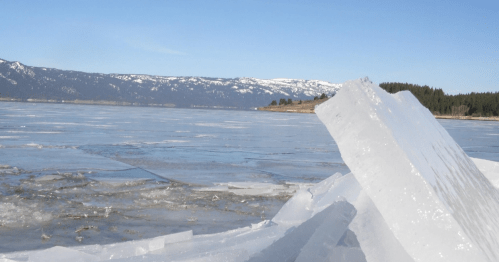 A frozen lake with large ice formations and snow-capped mountains in the background under a clear blue sky.