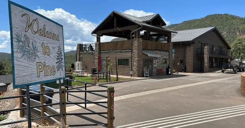 A welcome sign for Pine in front of a modern building surrounded by trees and mountains under a blue sky.