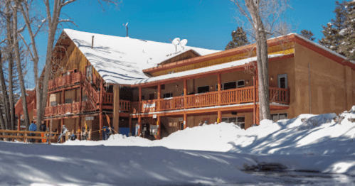 A rustic lodge surrounded by snow-covered trees, featuring wooden balconies and a sloped roof under a clear blue sky.