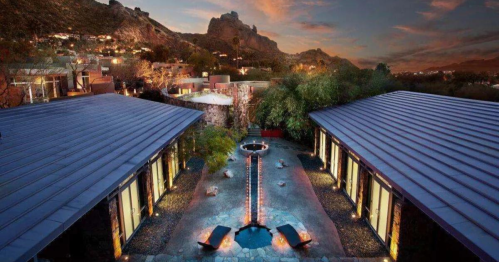 A serene outdoor space featuring modern architecture, a central fire pit, and a backdrop of mountains at sunset.