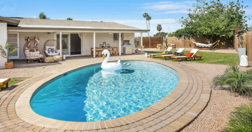 A sunny backyard with a pool, a swan float, lounge chairs, and a hammock surrounded by greenery.