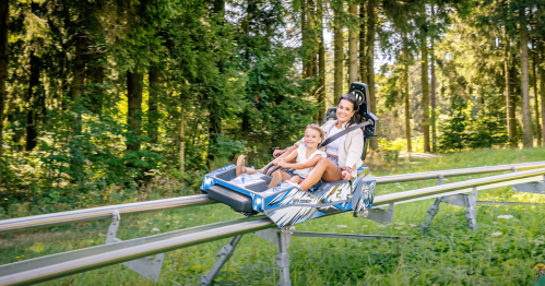 A woman and a child enjoy a thrilling ride on a mountain coaster through a lush green forest.