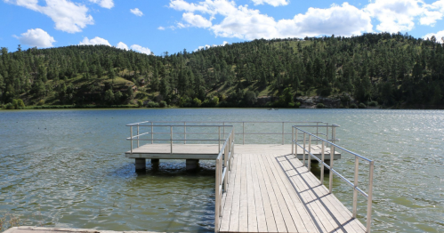 A wooden dock extends into a calm lake, surrounded by green hills and a blue sky with fluffy clouds.
