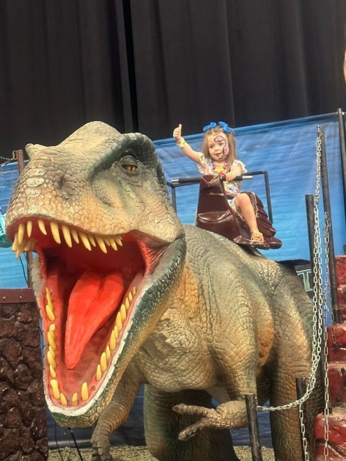 A child sits on a large dinosaur statue, giving a thumbs-up with a playful expression.