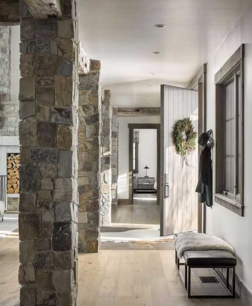 A modern hallway with stone columns, a wooden door, and a cozy bench, featuring natural light and a festive wreath.