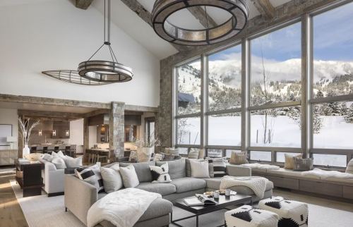 Modern living room with large windows showcasing a snowy landscape, featuring cozy furniture and rustic decor.
