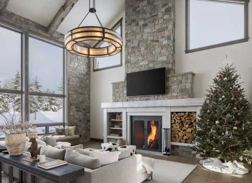 Cozy living room with a stone fireplace, large windows, a Christmas tree, and modern decor. Snow visible outside.