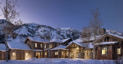 A cozy mountain lodge surrounded by snow-covered trees and mountains, illuminated at dusk.