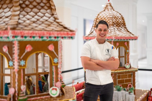 A chef stands proudly beside intricately designed gingerbread houses, showcasing his festive creations.