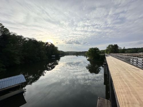 A serene river scene at sunset, reflecting clouds and trees, with a wooden walkway on the right.