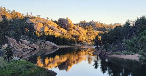 A serene lake surrounded by rocky hills and pine trees, reflecting the golden light of sunset.