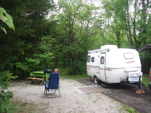 A small white camper parked at a campsite surrounded by lush green trees, with a picnic table and chairs nearby.