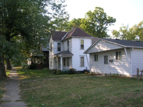 Two white houses with porches, surrounded by trees and grass, on a quiet street in a residential area.